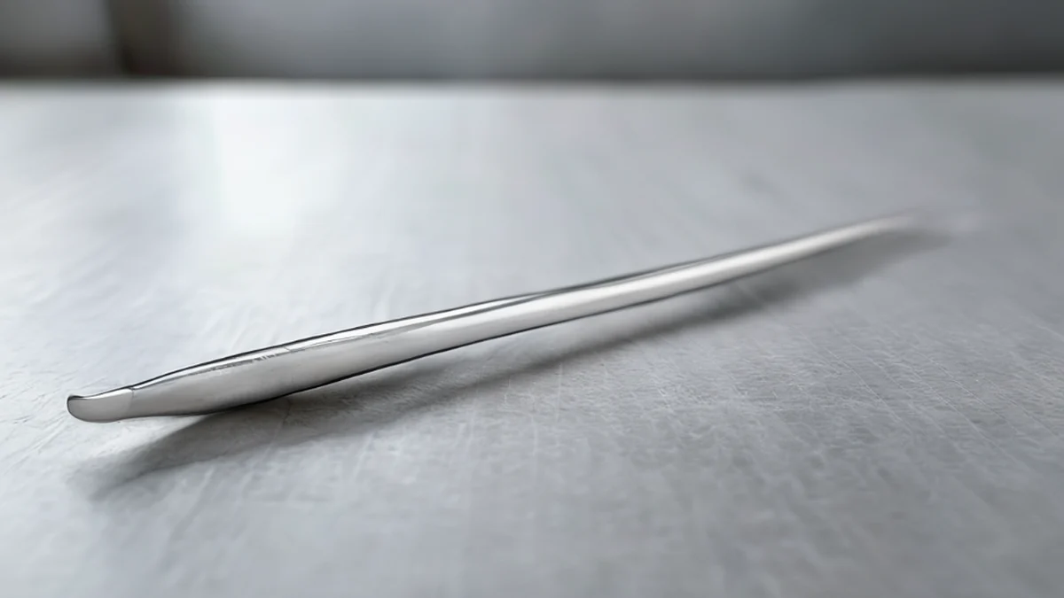 A single silver spindle, centered, with a long, thin shape, bright white color, and a smooth, polished surface, resting on a plain steel table, under overcast daylight, shallow depth of field.